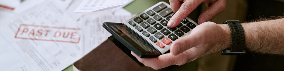 Senior Caucasian man using calculator while reviewing overdue bills, hands holding device and pressing buttons, past due notice visible on paperwork in background