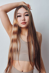 Naklejka premium Portrait of young woman with long straight hair wearing beige crop top posing with hand on head isolated on white background. Beauty, fashion and hairstyle concept with confident model.
