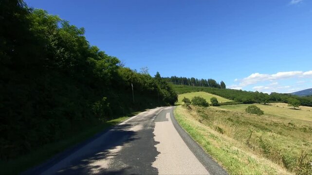 Scenic country road winding through lush green hills and forests in the Morvan region of France under a clear blue sky.