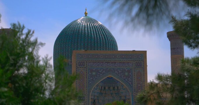 Cinematic video of turquoise tiled dome and ornate Islamic facade seen through soft foreground foliage clear blue sky detailed mosaic patterns calm heritage culture history travel and architectural