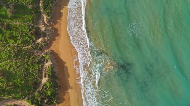 Picturesque tilt-down drone view of Riviera Beach at Għajn Tuffieħa, Malta, with turquoise Mediterranean waves washing over golden sand, bordered by lush green cliffs and a quiet coastal path.