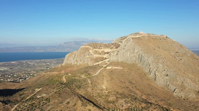 Aerial view of the Acrocorinth fortress atop a rocky hill with the surrounding Greek countryside and sea in the background under a clear blue sky.