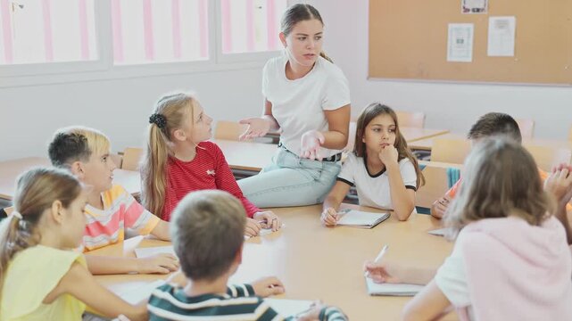 Friendly smiling teacher explaining studying material to diligent tweens sitting at common table and writing in notebooks during lesson at school