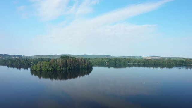 A tranquil view of Lac des Settons featuring a lush green forest island reflected on the still water under a blue sky. The peaceful natural landscape is surrounded by gentle hills and dense woodland.