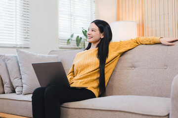 Fototapeta premium Happy young asian woman relaxing on comfortable sofa at home using laptop computer with yellow sweater looking away with cheerful smile