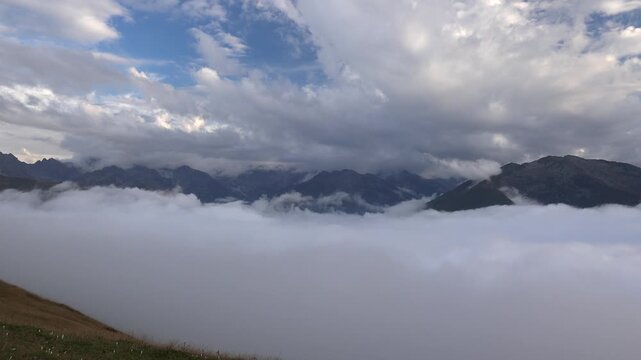 Mountain valley blanketed by low clouds above alpine meadow, dramatic sky and peaks. Misty ridge scene with overcast weather, rolling fog layer, remote highlands, rugged summits.