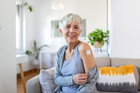 Happy senior woman showing a bandage on her arm after vaccination. Smiling mature lady with grey hair sitting on a sofa after getting a vaccine shot. Healthcare, immunization and flu shot concept.