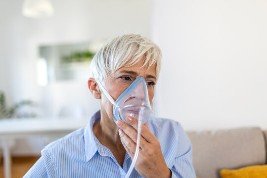 Senior woman using an oxygen mask for respiratory therapy at home. Mature lady with short grey hair holding a nebulizer mask. Healthcare, asthma, pneumonia and breathing problem medicine concept.