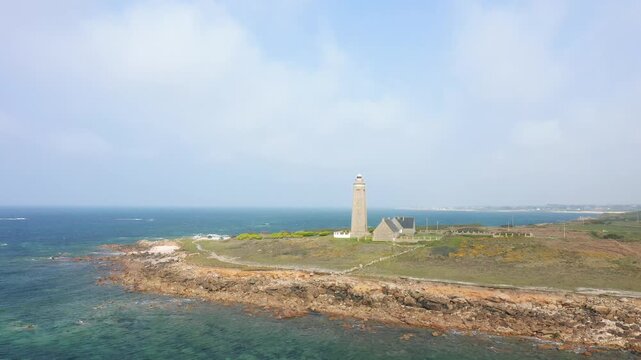 Aerial view of the Cap Levi Lighthouse standing on a rugged coastline with clear blue sea in Normandy, France. The scene features dramatic rocks, coastal vegetation, and expansive ocean under a partly