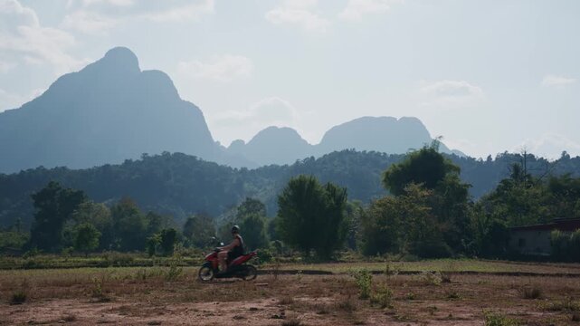 Tourist on a scooter explores Laos countryside with majestic limestone mountains in background during a sunny day