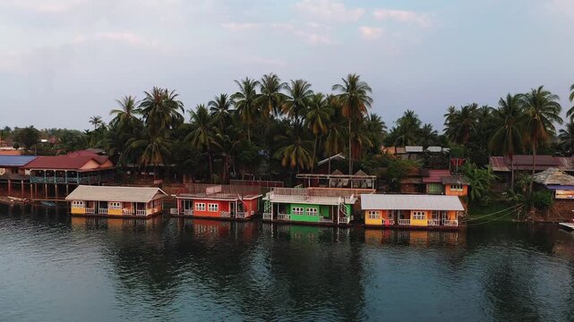 A row of vibrant wooden houses on stilts lines the riverbank, framed by lush palm trees in the 4000 Islands region of the Mekong. Tranquil water reflects the colorful facades under a soft sky.