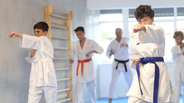 Concentrated teenager in white kimono practicing punches in gym during group martial arts workout. Shadow fight, combat sports training concept 