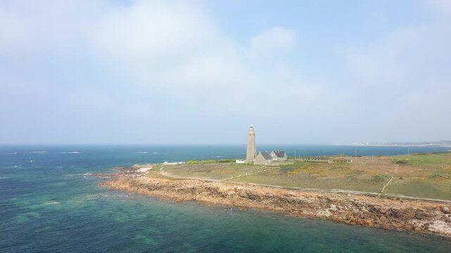 Aerial view of the Phare du Cap Levi lighthouse standing on a rugged coastline in Normandy, France, with the blue sea in the background. Tranquil seascape and open sky create a serene coastal scene.
