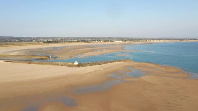 Aerial view of the expansive sandy beach and calm blue waters at Grande Plage de Barneville, Normandy, France. The scene features tidal pools, breakwaters, and a distant town under clear skies.