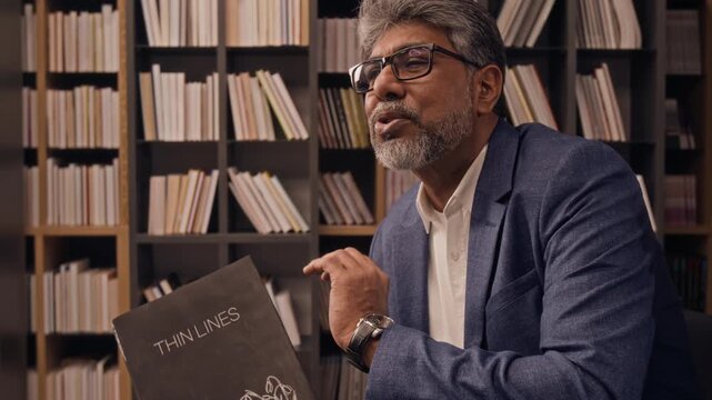 Low angle tilt-up shot of middle-aged biracial bearded author with glasses talking about his new book, holding it in his hands in library