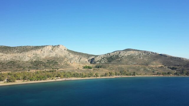 Aerial view of Paralia Karathona beach with clear blue water, sandy shoreline, and rocky hills in Nafplio, Greece under a clear sky.