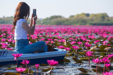 A young woman sitting on a boat is using her mobile phone to take pictures of the red lotus lake.