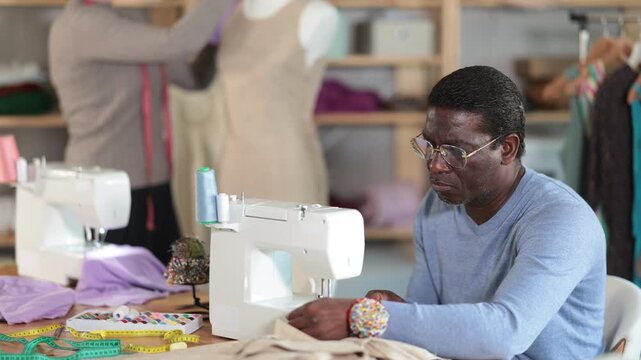 African American man tailor sews clothes in sewing machine. Worker uses sewing machine to create clothes for clients. Companion work in background with mannequin