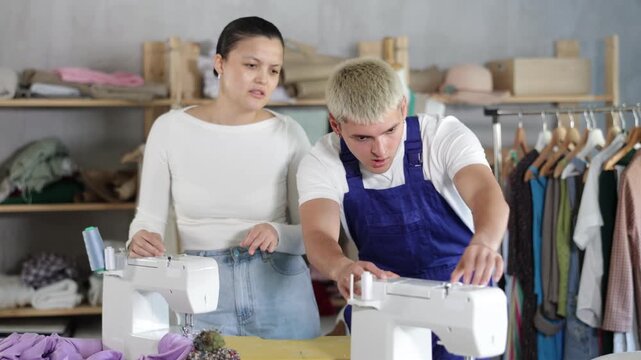 Focused young technician fixing stitching-machine while anxious dressmaker explains issue beside him in sewing studio
