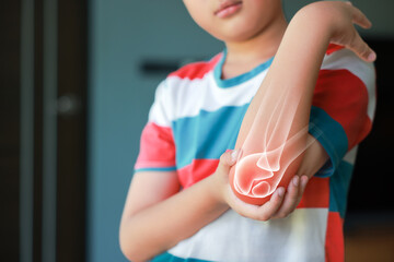 Fototapeta premium Little boy suffering from elbow joint pain with red x-ray bone graphic overlay, child touching arm feeling ache from injury, inflammation, fracture or accidental fall while playing at home.
