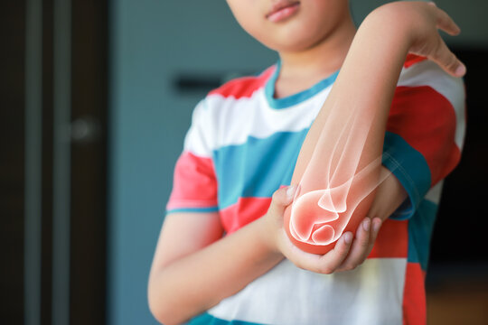 Little boy suffering from elbow joint pain with red x-ray bone graphic overlay, child touching arm feeling ache from injury, inflammation, fracture or accidental fall while playing at home.