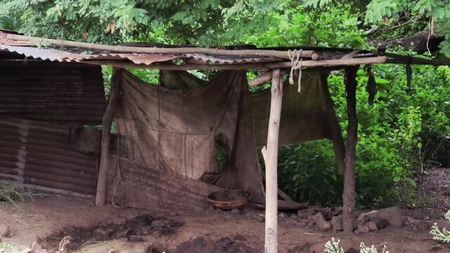 A rustic Indian village cattle shed with a corrugated metal roof and wooden posts. Fabric curtains offer basic shelter for livestock, an empty dung bowl rests on the dirt floor of open-air structure.