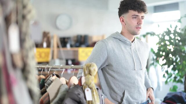  Regular customer guy client pre-examine fabric and style of product, is busy choosing jeans trousers. Client enjoys shopping for clothes at store browsing through colorful on display racks
