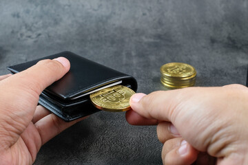 A hand inserts a bitcoin coin into a black wallet on a dark surface with coin stacks nearby