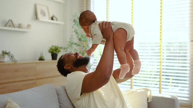 Happy African American father lifting his baby high in the air while sitting on a couch in a bright living room. Joyful bonding moment and parenthood concept in a sunlit modern home interior.