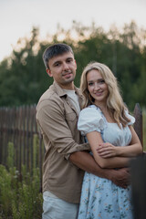 Obraz premium A young Caucasian couple stands together in a field. The man has short gray hair and wears a beige shirt. The woman has long blonde hair and a floral dress.