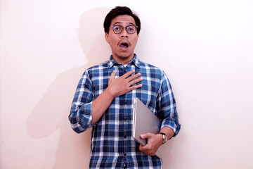 Fototapeta premium Southeast Asian man wearing flannel shirt standing and holding laptop with shocked expression, on white isolated background.