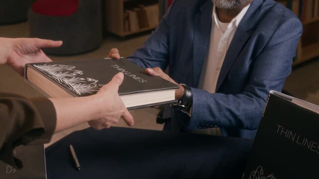 High angle shot of unrecognizable man in formal outfit selling copies of his books while sitting at table in book store