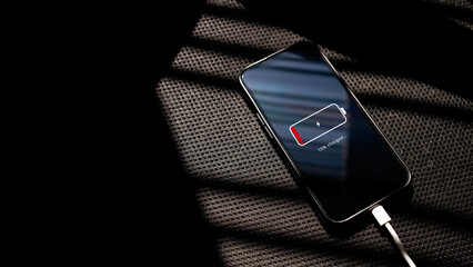 Smartphone with low battery 10% charging via cable on a black chair textured surface. Concept representing power shortage, energy management