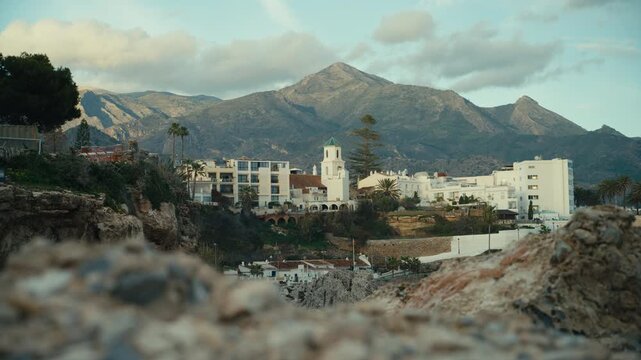 Telephoto view of Nerja&rsquo;s whitewashed old town with church tower and cliffside buildings framed by blurred rocks in foreground and dramatic Andalusian mountains behind.