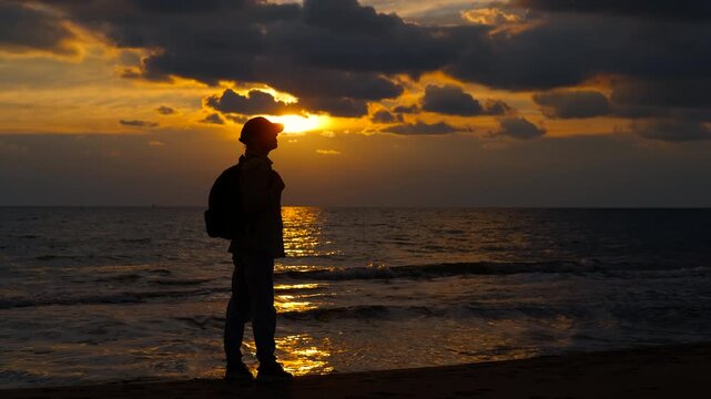 Silhouette of traveler enjoying the sunset on the beach. Solitary traveler watching a dramatic golden sunset over the ocean. The man stands on the sandy beach, observing the sun e