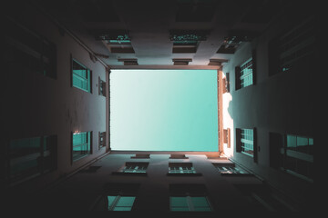 Looking straight up through a dark narrow courtyard, tall apartment walls frame a clean turquoise sky like a glowing minimalist geometric portal   © Marci