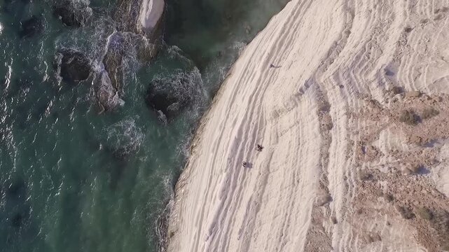 Aerial side view of Scala dei Turchi white cliffs rising from turquoise sea waves, Sicily, Italy, layered rock face with distant beachgoers and rocky outcrops.