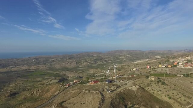 Aerial view of wind turbines on a hill overlooking rural landscape near Agrigento, Sicily, Italy, with Mediterranean Sea, fields and distant village under clear blue sky.