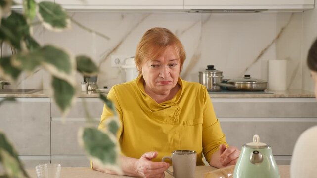 Elderly woman in casual clothes is outraged by communication with her daughter-in-law. Women talk about nasty things in kitchen at table and drink tea or coffee