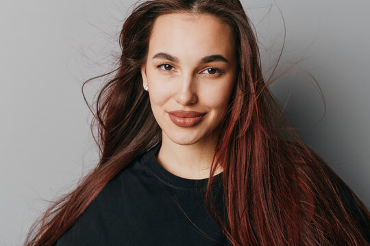 Young woman with long hair and natural makeup smiling at camera on gray background. Female portrait with casual black top and confident expression for beauty and lifestyle.