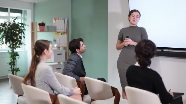 Positive young girl coach standing near interactive board telling new topic or showing presentation with remote control to people sitting on chairs in light classroom