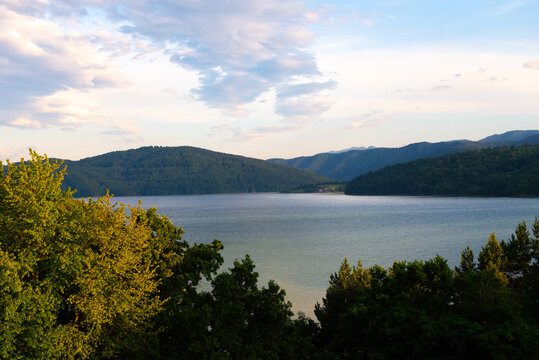 A peaceful mountain scene featuring a calm lake surrounded by lush greenery and towering hills, illuminated by soft light during the golden hour.