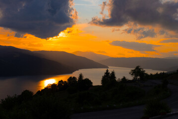 Fototapeta premium A stunning sunset casts golden hues over a calm lake surrounded by mountains. The sky is filled with clouds, creating a breathtaking view of nature's beauty.