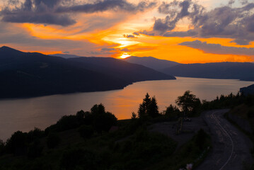 Obraz premium The sun sets behind the mountains, casting golden hues over the calm lake. Trees line the shore, creating a peaceful and picturesque mountain scene.
