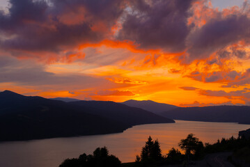 Naklejka premium A vibrant sunset casts warm colors over a serene lake nestled between towering mountains, creating a peaceful atmosphere in nature's embrace.