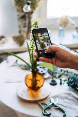 Hand holds smartphone to photograph floral arrangement in glass vase on table. influencer lifestyle, floral blogging, behind the scenes, mobile photography, sharing hobbies online