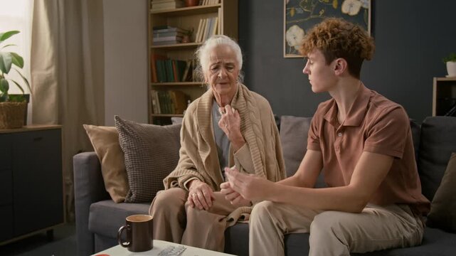 Curly-haired teenager giving digital thermometer to sick grandmother who putting it under armpit and coughing, grandson patting her on back trying to reassure