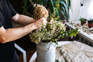 Hands style delicate hydrangea and green foliage in white vintage milk can on table. Ethereal flower arrangement, romantic cottagecore bouquet, literary floral aesthetic