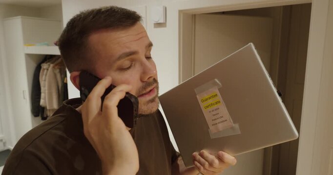 Man calling customer service while holding a defective laptop with guarantee certificate attached. Concept of warranty repair request, consumer protection, technical issue and after-sales service.