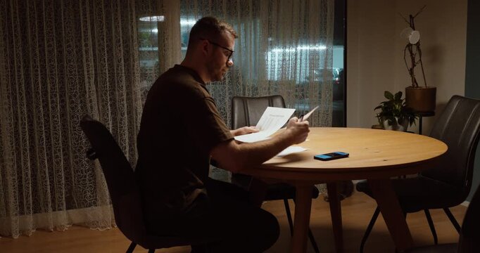Adult sitting at dining table in the evening reviewing mortgage documents with concerned expression. Concept of financial stress, rising interest rates, debt burden, housing costs and economic